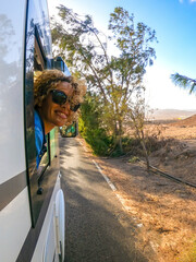 Blonde woman with curly hair looks out of the window of the moving camper. Female tastes the wind in her face and smiles. Travel and tourism concept.