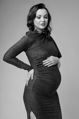 Black and white portrait of young pregnant female in grey sequin dress with hands near pregnant belly. - 787710511
