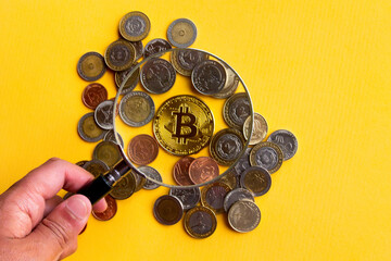 a hand with a magnifying glass focusing on the cryptocurrency bitcoin among several Argentine currencies on a yellow background, concept of inflationary crisis and search for solutions on the web3