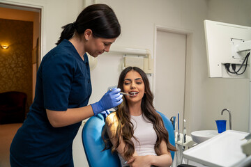 Young woman with dental braces having in dental office , wide shot, open mouth, female dentist using mirror for checkup 