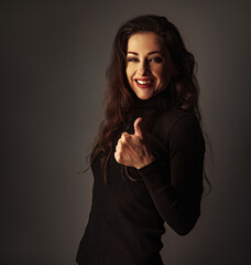 Beautiful smiling business woman in black clothing looking happy and showing thumb up success sign by hand on dark shadow studio background with empty space. Closeup