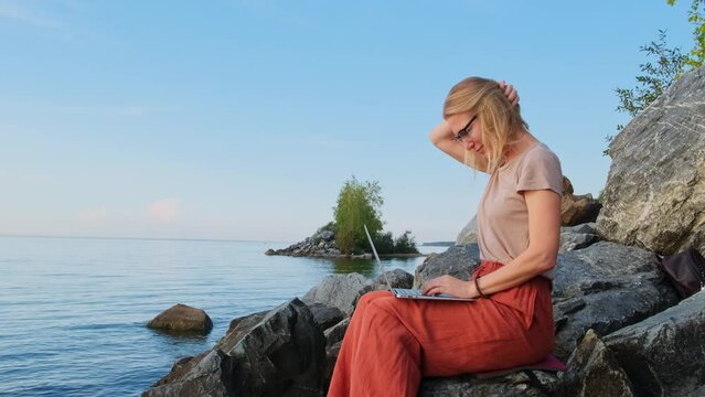 A digital nomad, blonde caucasian woman sits by the sea and works remotely. Female freelancer uses laptop computer on empty seashore. Office for introvert outdoor.