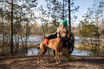Middle aged woman petting Hungarian Vizsla dog walking in nature autumn park. Female travelling...
