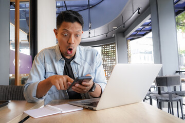 Asian man shocked face, wow, surprised with blue shirt using laptop and mobile phone in coffee shop, shopping online - obrazy, fototapety, plakaty