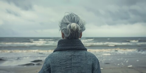   A woman with her back to the camera faces a body of water on the beach - obrazy, fototapety, plakaty