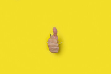 Hand with thumb up as a sign of approval, coming out of the hole of a yellow torn paper background. - obrazy, fototapety, plakaty