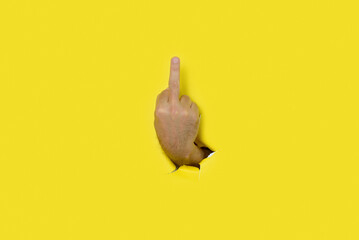 Hand of man making insulting gesture with middle finger through a hole in yellow paper background....