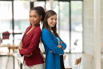 Two diverse businesswomen standing back to back with arms crossed, smiling confidently in a bright modern office, conveying partnership, leadership, and empowerment