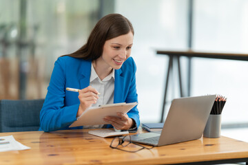 Smiling businesswoman working remotely, participating in an online webinar or video conference from her home office, taking important notes on a clipboard while looking at her laptop screen