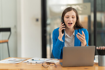 Young businesswoman managing business communications while talking on a smartphone and working on a laptop at a desk, embodying professional multitasking and efficiency in a modern office setup