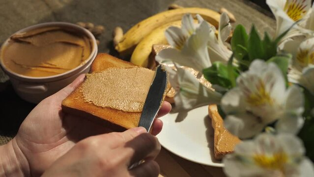 A woman uses a table knife to spread peanut butter on crusty white toast. The hand holding the bread applies a layer of nut paste using a knife. High-calorie breakfast made from natural ingredients