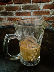 The remaining coffee drink is in a clear glass cup on a glass table with a brick wall as a background, taken wide at eye level