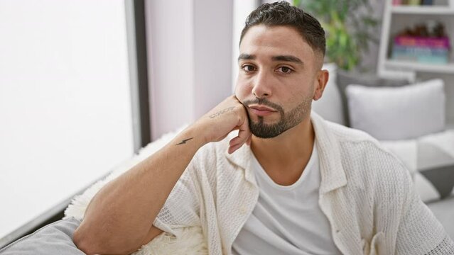 Serious and trusting young arabian man with beard, perfectly capturing a natural simple expression as he sits on his home sofa, looking at the camera, indoor lifestyle becoming his style