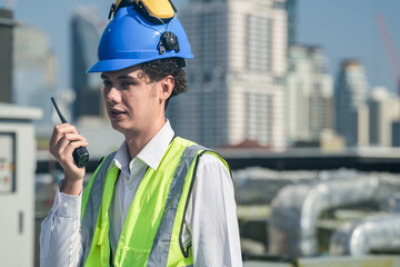 Focused engineer in hard hat and safety vest using walkie-talkie and laptop on construction site...