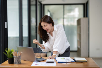 Joyful businesswoman with a victorious gesture, celebrating success while working on her laptop in a sunny office space.