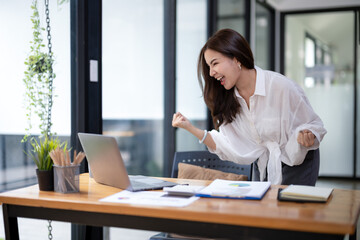 Joyful businesswoman with a victorious gesture, celebrating success while working on her laptop in a sunny office space. - obrazy, fototapety, plakaty