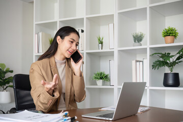 Young Asian business woman sits on the phone in an online business meeting using a laptop in a modern home office decorated with shady green plants.