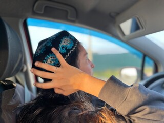 Brunette Uzbek girl looking in the rearview mirror while touching national headwear of Central...