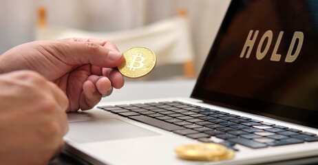 A person is holding a bitcoin gold coin on a laptop screen that says "Hold"
