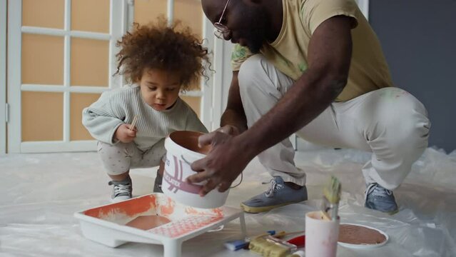 Handheld medium footage of Biracial little girl watching her African American dad pour beige paint from bucket into tray before painting walls in new apartment