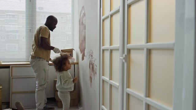 Side full footage of single African American dad painting wall and renovating nursery room for his cute little daughter who helping him