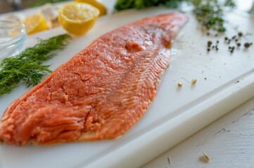 Raw half salmon fillet with ingredients such as herbs, lemon, salt and pepper on a white cutting board