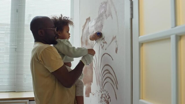 Side footage of African American man holding his little Biracial daughter in hands and teaching her how to paint walls with roller during room redecoration