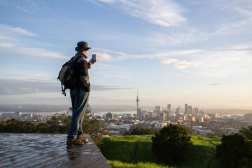Tourist taking photos of Sky Tower and cityscape using smartphone. Mt Eden summit at sunrise. Auckland. - 787949307