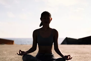 Tuinposter backlight of young woman doing meditation by the sea sitting with legs crossed, concept of mental relaxation and healthy lifestyle © Raul Mellado