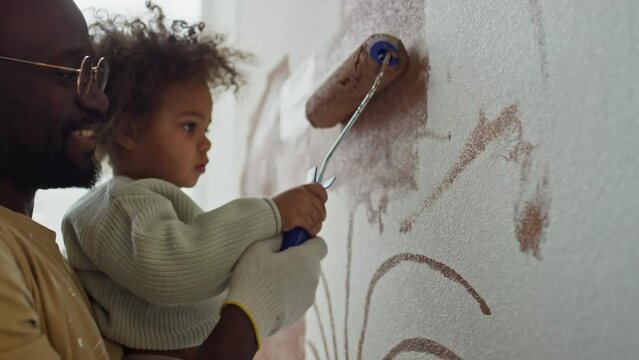 Side closeup of positive African American dad holding his Biracial baby daughter in hands and helping her to paint wall in brown using roller