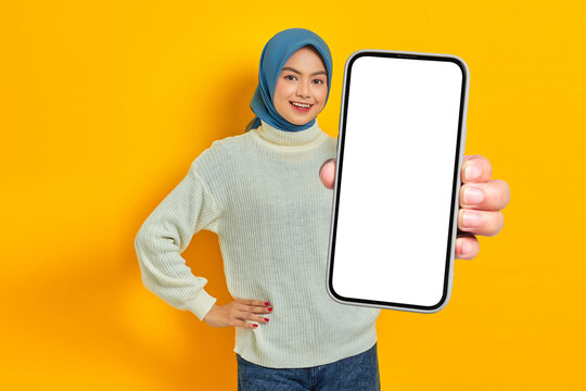 Smiling beautiful Asian woman in white sweater and hijab, showing blank mobile phone screen isolated on yellow background. Muslim lifestyle concept