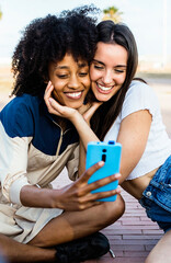 Vertical photo of young happy multiracial women friends taking a selfie with smartphone app sitting outdoors in summer. LGBT lesbian couple taking self photography. Female friendship concept. 