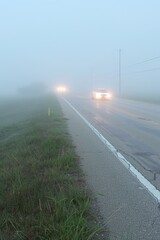 Misty, oppressive clouds shroud a lonesome highway, sporadic headlights cutting through fog, the road gleaming