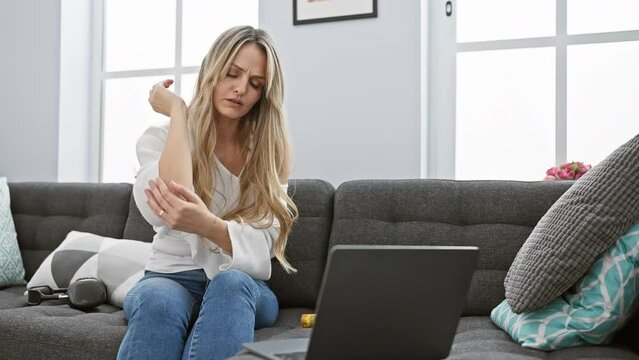 A young blonde woman suffering from elbow pain while sitting with a laptop and medication on a couch indoors.
