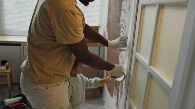 Side tilt footage of Black man sticking masking tape along doorway during paintwork in future nursery, baby helping him and painting wall with mini roller