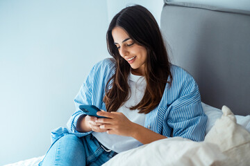 Young woman sitting on bed, dressed in casual clothes, holding phone and reading new messages,...