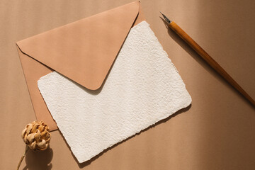 Overhead view of blank piece of homemade deckle edge card, envelope, flower and calligraphy pen on a wooden table