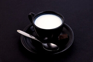 Black cup with milk on a saucer with a spoon and a bar of chocolate on a black table.