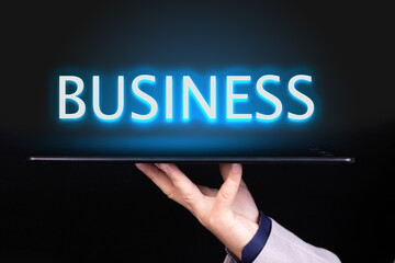Man hand holds a tablet over which a neon text is written, the word BUSINESS. Business concept.