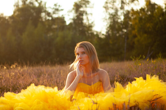 Amidst a field at sunset, a woman sits pensively, her voluminous yellow gown billowing around her. The golden hour light softly illuminates her thoughtful expression, and the natural backdrop fades