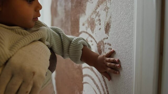 Side chest up footage of Biracial baby girl creating handprints on wall with brown paint during nursery redecoration, African American dad holding her in hands and smiling