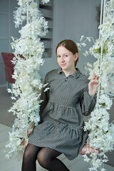 Portrait of a beautiful young girl sitting on a swing in a photo studio. Floral light interior - 788033379