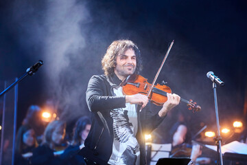 A violinist musician stands on stage with a violin in his hands during a concert. There is a bright light from the floodlights around. - 788035544