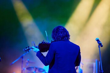 A violinist musician stands on stage with a violin in his hands during a concert. There is a bright light from the floodlights around. - 788035545
