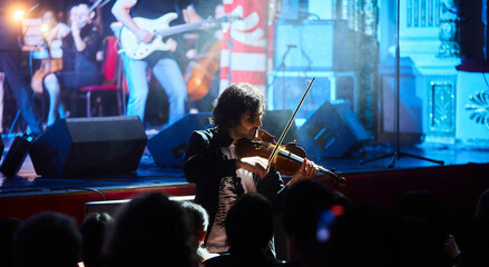 A violinist musician stands on stage with a violin in his hands during a concert. There is a bright light from the floodlights around. - 788035549