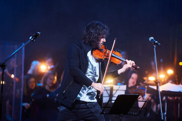 A violinist musician stands on stage with a violin in his hands during a concert. There is a bright light from the floodlights around. - 788035564