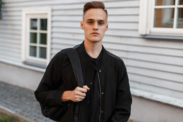 Cool man model with a hairstyle in a black bomber jacket with a backpack is walking on the street...