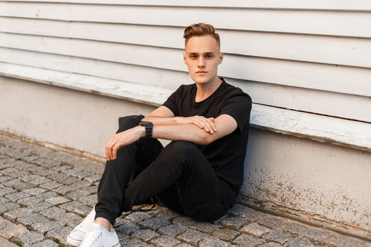 handsome fashion man model with a hairstyle in black clothes with a t-shirt and jeans with white sneakers and a backpack sits near a wooden house