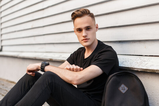 Handsome youth man model with hairstyle in a black T-shirt with a backpack sits on the street near a wooden wall