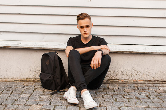 young handsome man with a hairstyle in a black T-shirt with jeans and white sneakers with a backpack sits near a wooden building